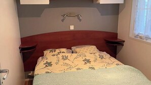 A bedroom with a bed, two nightstands, a wooden headboard, a built-in shelf above the bed, and a window with curtains. - Mobile home for 6 people. (Arvert)