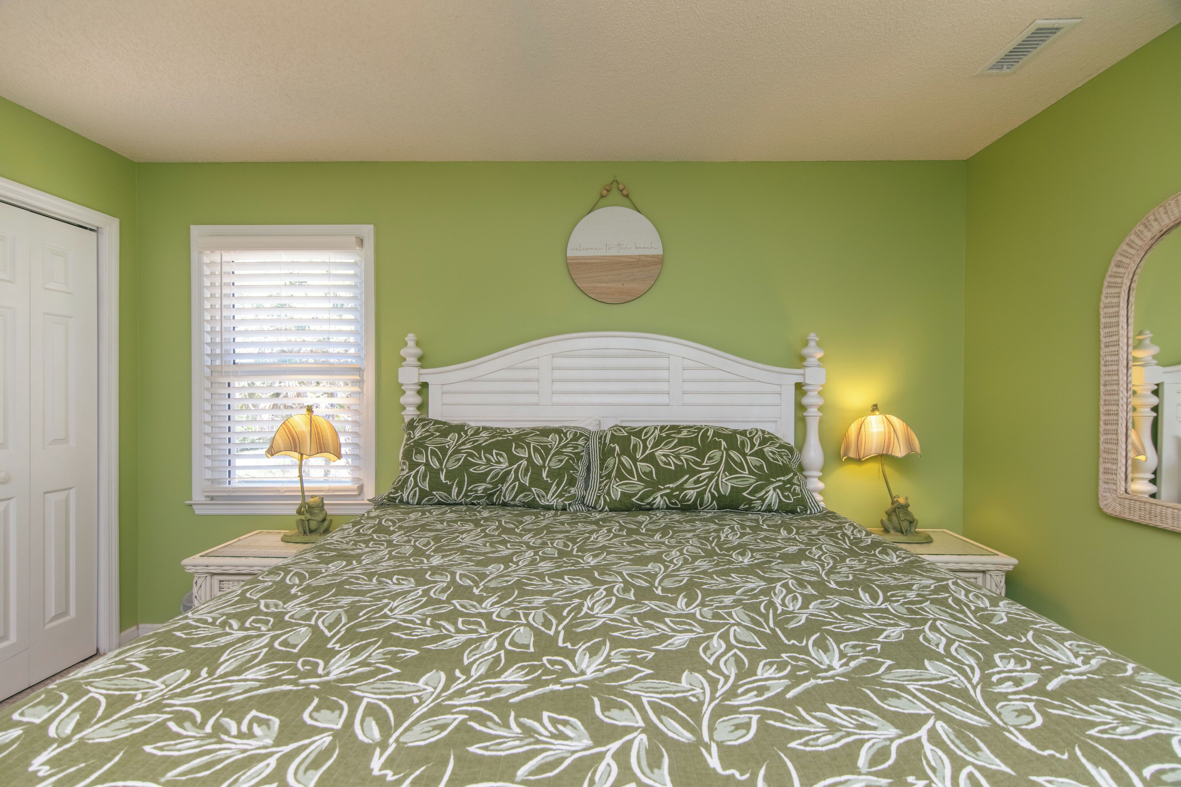 A bedroom with a bed, two bedside lamps, a window with blinds, a door, and a mirror on the wall.