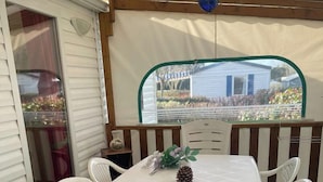 En täckt uteplats med ett bord och stolar, en hängande lampa och utsikt över ett hus genom ett fönster.