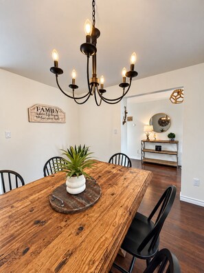Un coin repas avec une table en bois, des chaises noires, un lustre et un panneau accroché au mur.