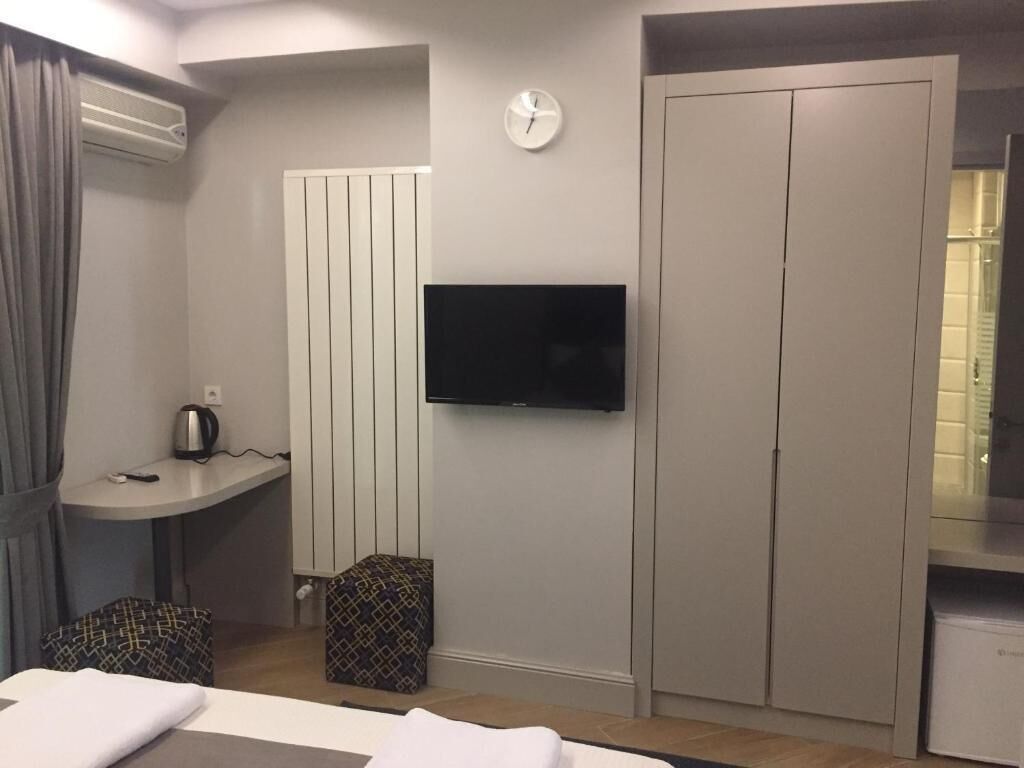 A hotel room with a bed, a television mounted on the wall, a wardrobe, a small table with a kettle, and a window with curtains.