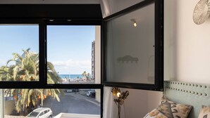 A hotel room with a large window overlooking a beach, a bed with patterned pillows, and a decorative wall lamp.