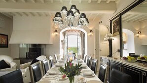 Une salle à manger avec une longue table dressée pour un repas, un lustre et une vue sur l’extérieur par une porte ouverte.