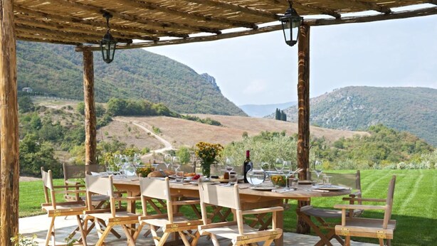 Ein Essbereich im Freien mit einer Holz-Pergola, gedeckten Tischen und einem schönen Blick auf sanfte Hügel im Hintergrund.