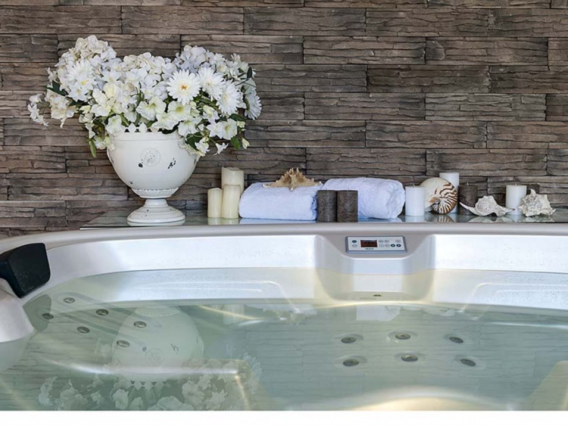A spa area with a hot tub, a decorative vase with white flowers, candles, and a stone wall backdrop.