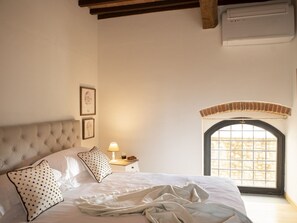 A bedroom with a tufted headboard, a bed with white linens, a nightstand with a lamp, and a window with a view of stone walls. - Luxury refined villa with pool (Tavarnelle Val di Pesa)