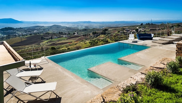 A rectangular swimming pool with clear blue water, surrounded by a concrete deck. There are lounge chairs and a small table on the deck. The pool is set against a backdrop of rolling hills and distant mountains.