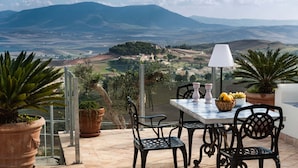 A terrace with a table and chairs, potted plants, and a view of a valley with mountains in the background.