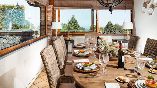 A dining area with a wooden table set for a meal, wicker chairs, and a view of greenery through large windows. - Luxurious villa on the Etna slopes, with private pool and fitness area (Viagrande)