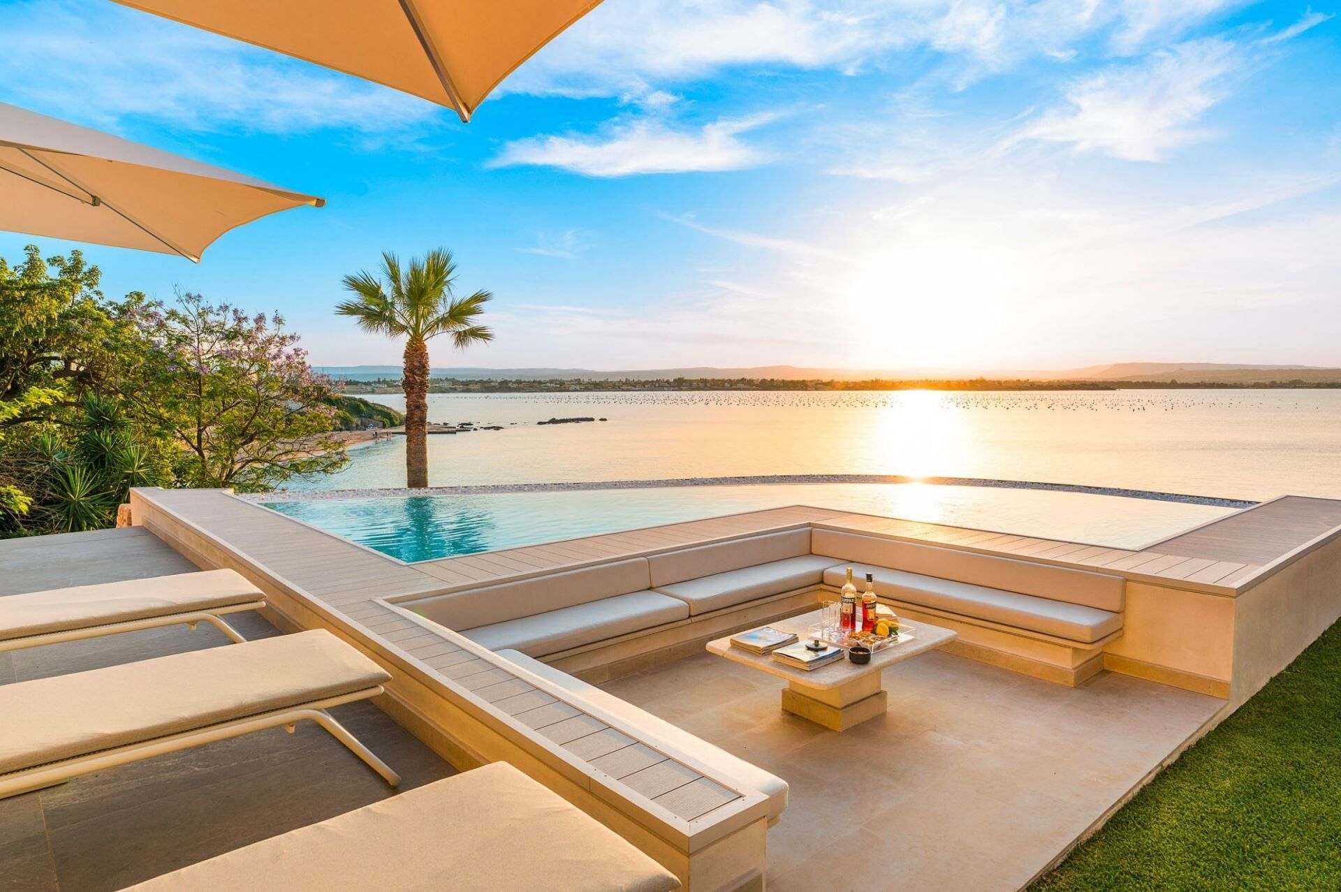 A poolside area with a view of the sunset over the water, a palm tree, and outdoor seating.