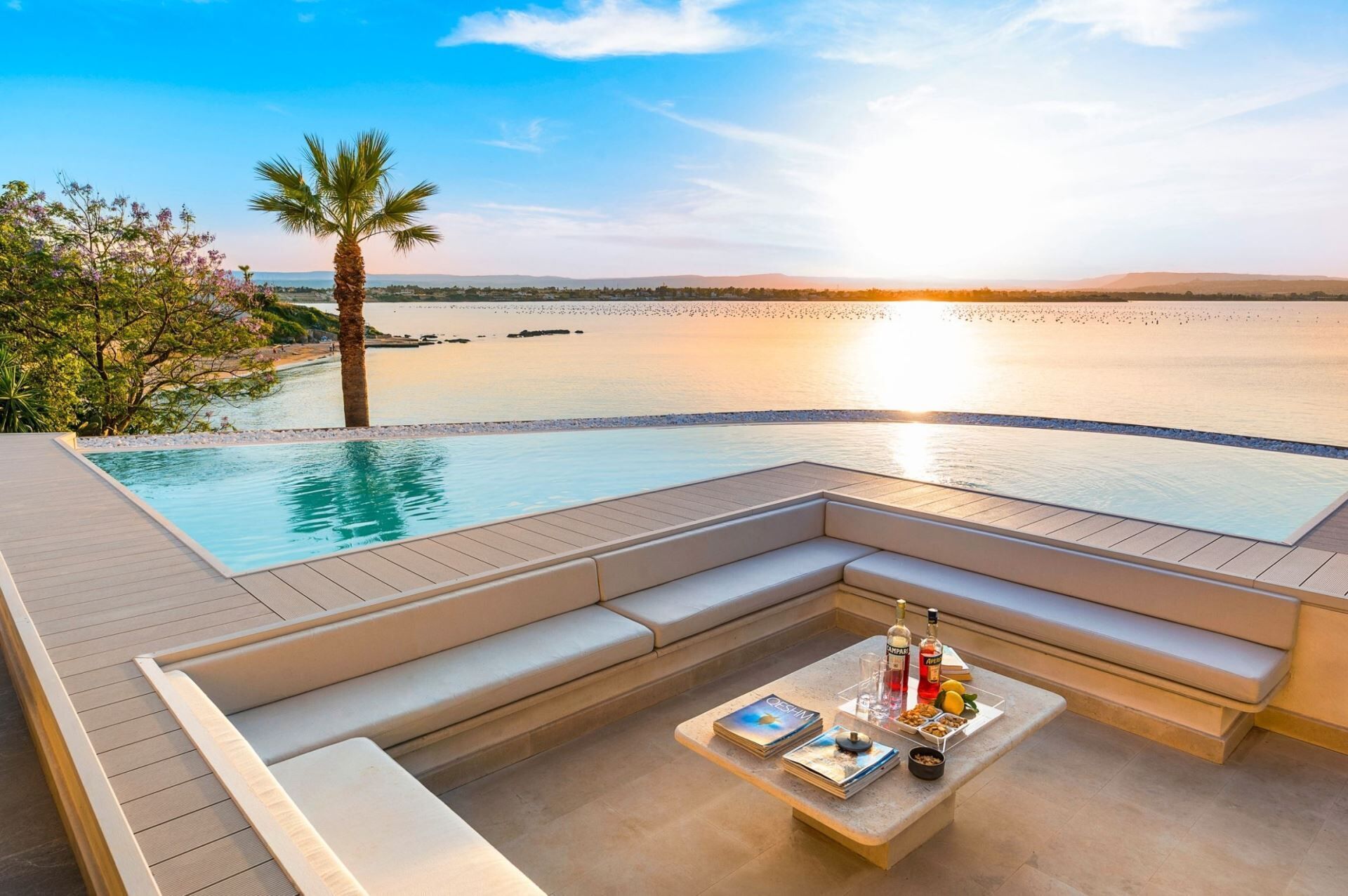 A poolside area with a view of the sea at sunset, a palm tree, and a seating area with a table and drinks.