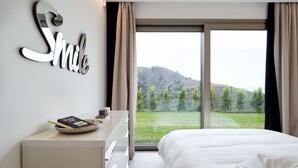 A modern hotel room with a large bed, a bedside table with a book and a remote control, a wall-mounted mirror, and a view of a green landscape through a large window. - Dream Villa with heated pool and magnificent view on the Taormina Bay (Letoianni)