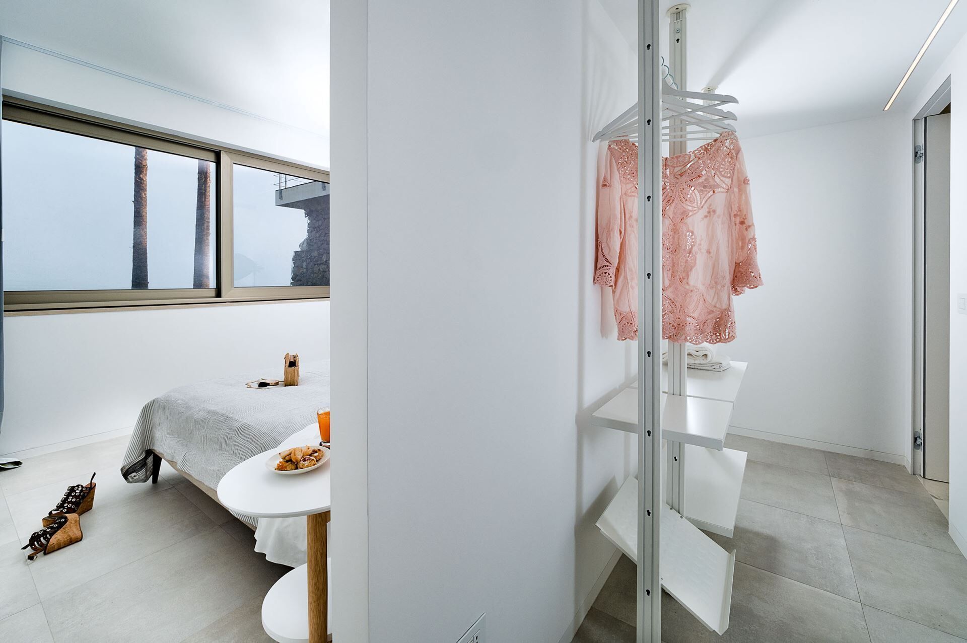A modern, minimalist bedroom with a large window, a bed, a small table with a plate of food, and a white wardrobe with a hanging rack.