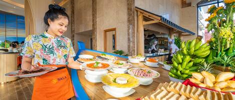 Een buffetopstelling met diverse gerechten en fruit, een bedienend personeelslid in een schort, en een modern interieur met grote ramen en een hoge plafond.