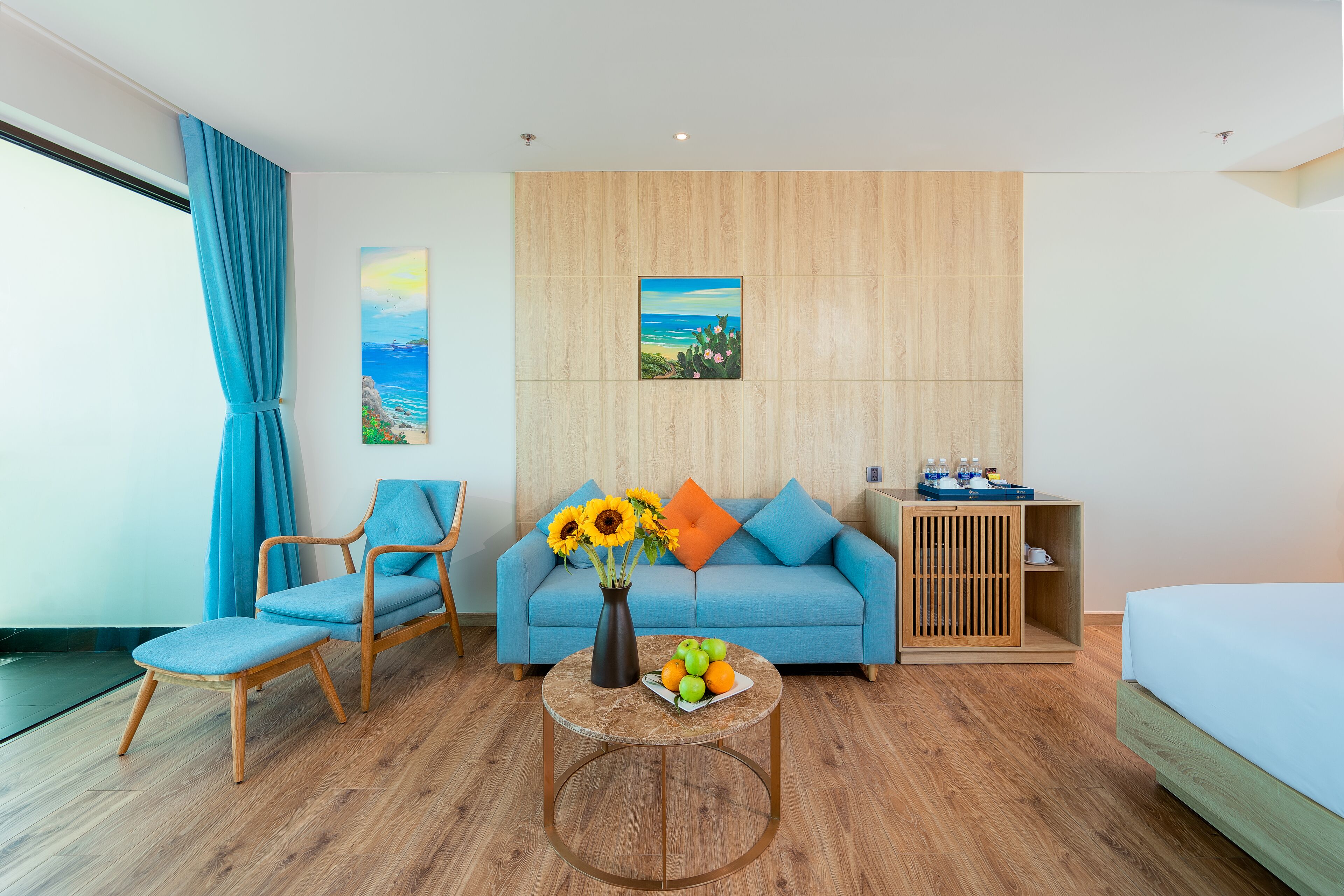 Une chambre d’hôtel moderne avec un plancher en bois, un canapé bleu, une chaise en bois, une petite table ronde ornée d’un vase de fleurs et un tableau accroché au mur.