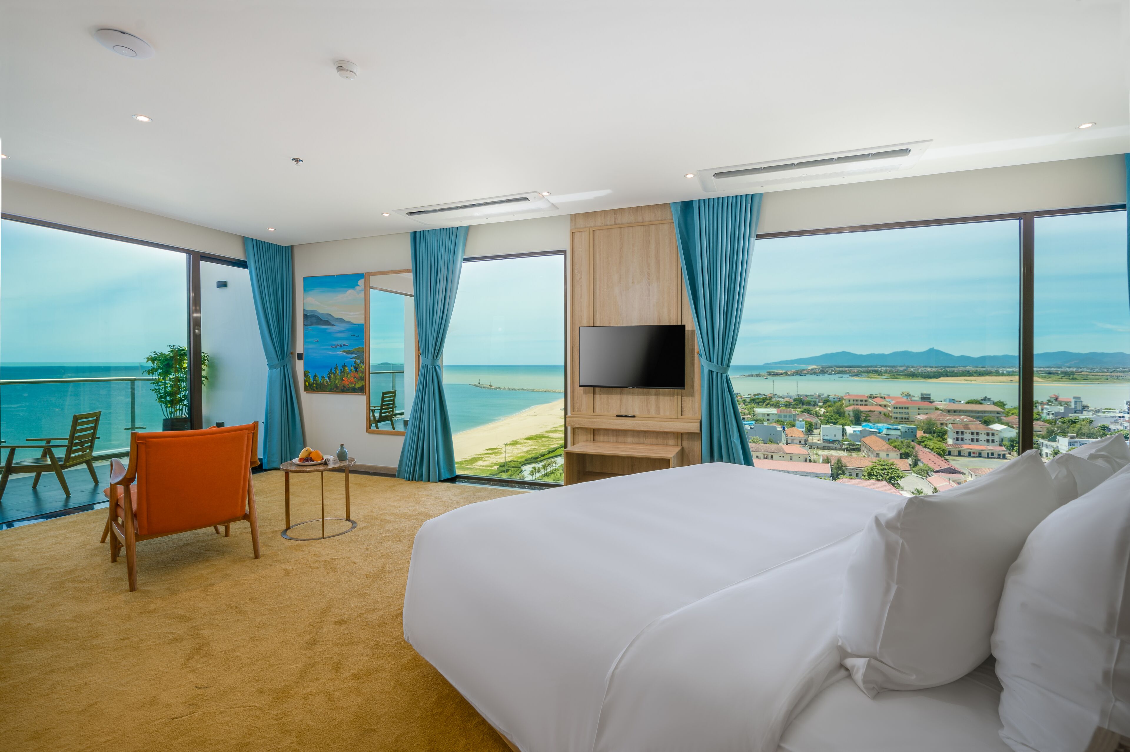 A hotel room with a large bed, a TV, a desk, and a view of the beach and city.