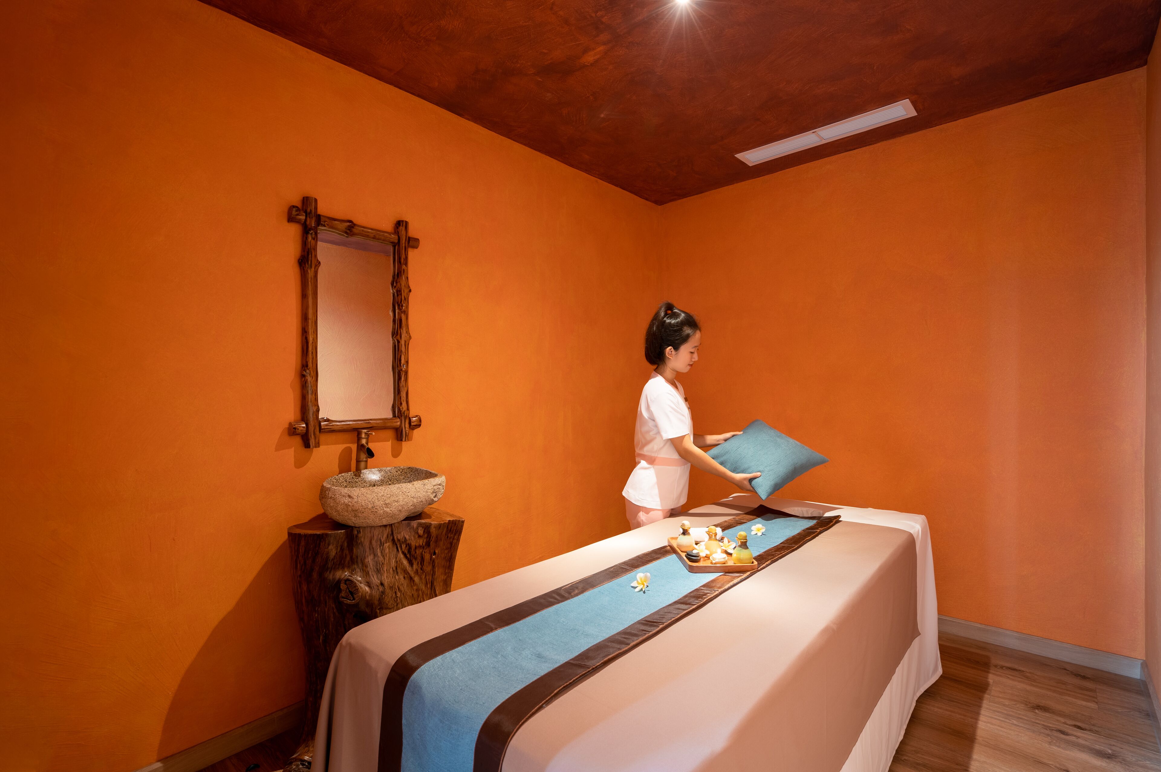 A spa room with an orange wall, a wooden basin, a bed with a blue bed runner, and a mirror.