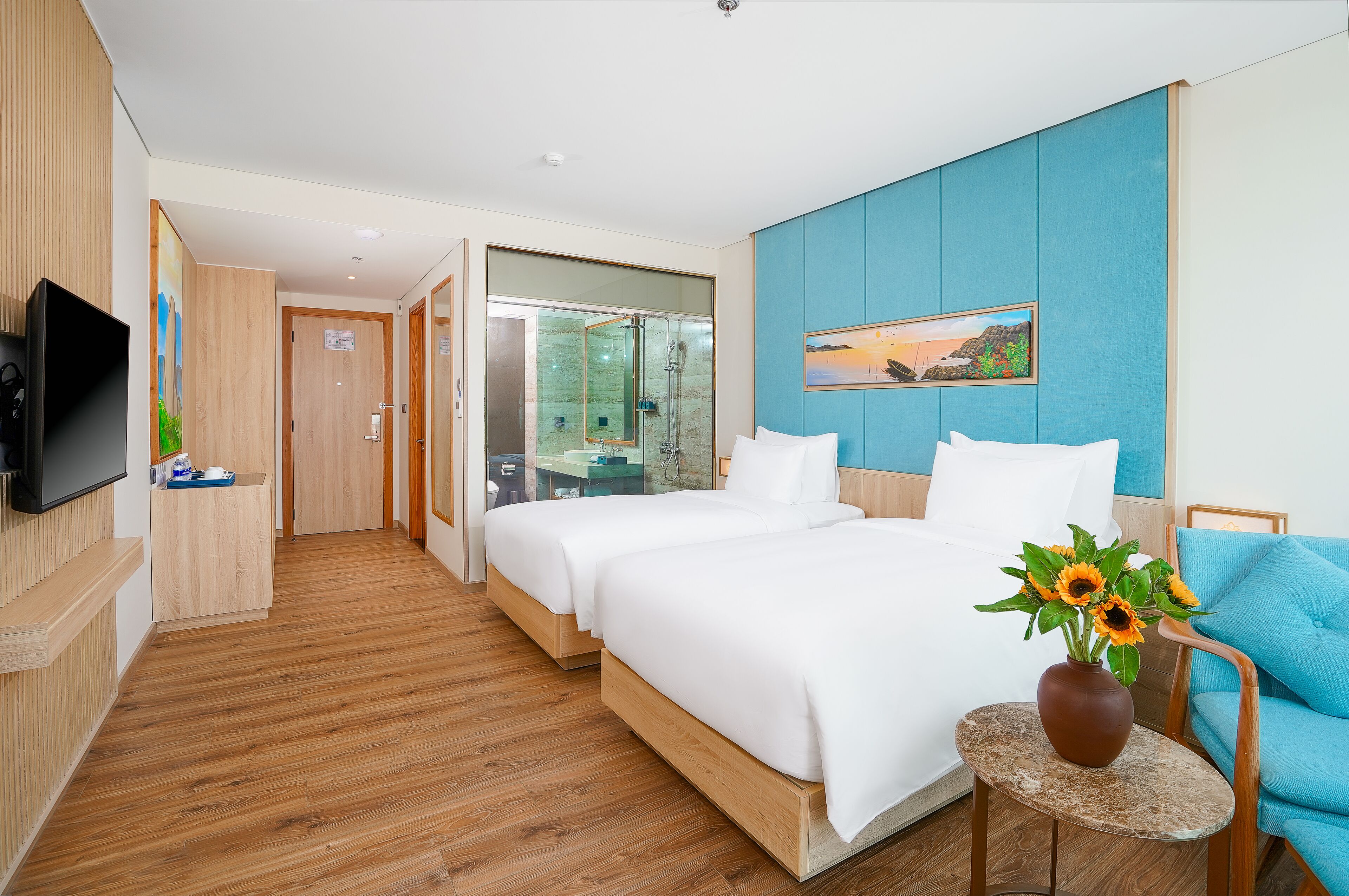 Een hotelkamer met twee bedden, een televisie, een klein tafeltje met een bloevas en een blauwe fauteuil.