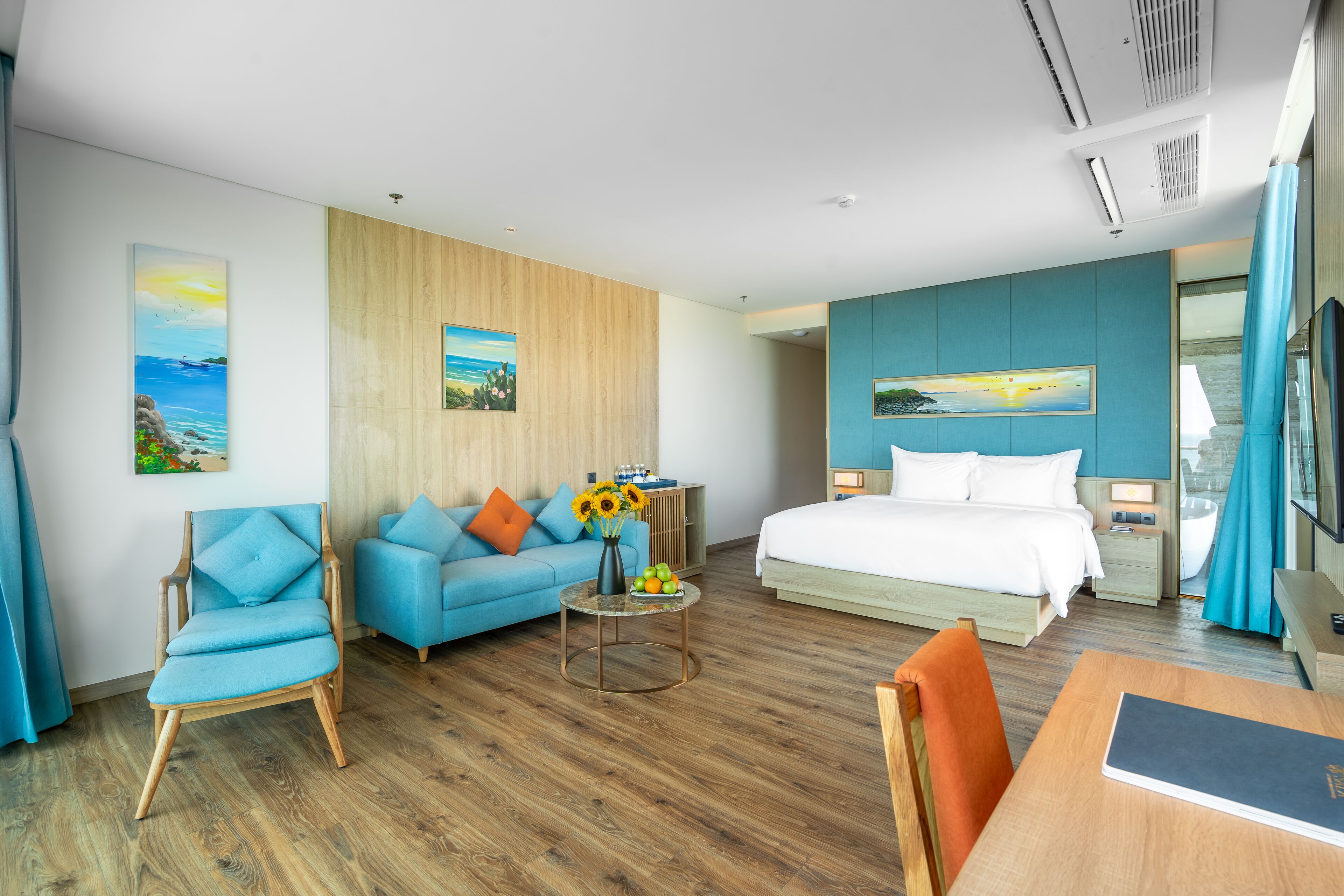 A modern hotel room with a wooden floor, a bed with white linens, a blue sofa, a small round table with a vase of flowers, and a desk with a chair.