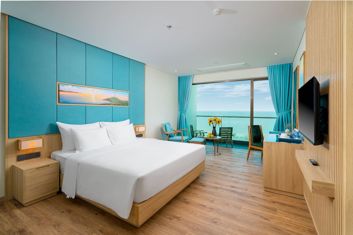 Ein Hotelzimmer mit einem großen Bett, einem Schreibtisch mit Flachbildfernseher, einem Essbereich mit Tisch und Stühlen sowie einem großen Fenster mit Vorhängen.