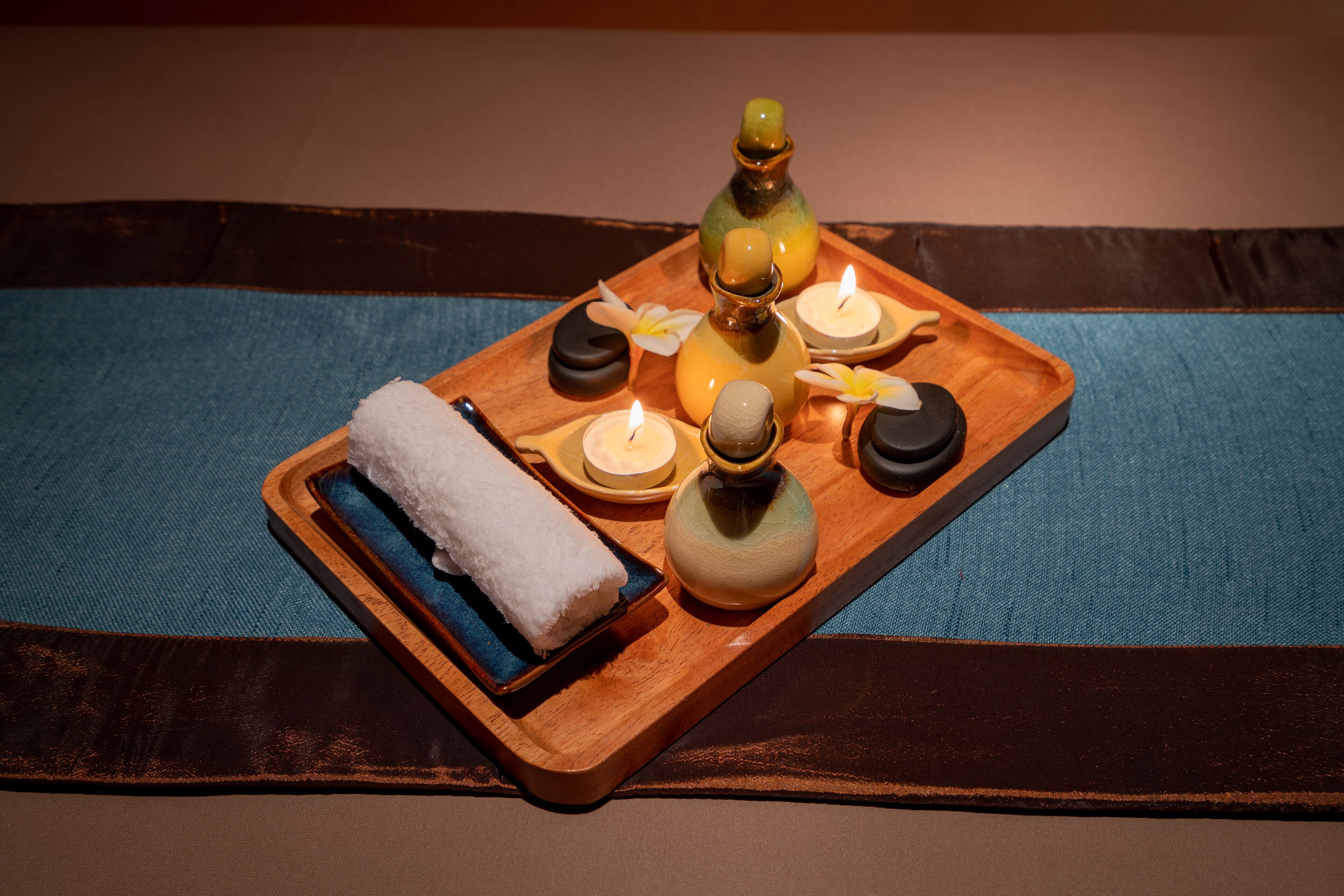 A wooden tray with candles, a bottle, and a towel on a blue fabric surface.