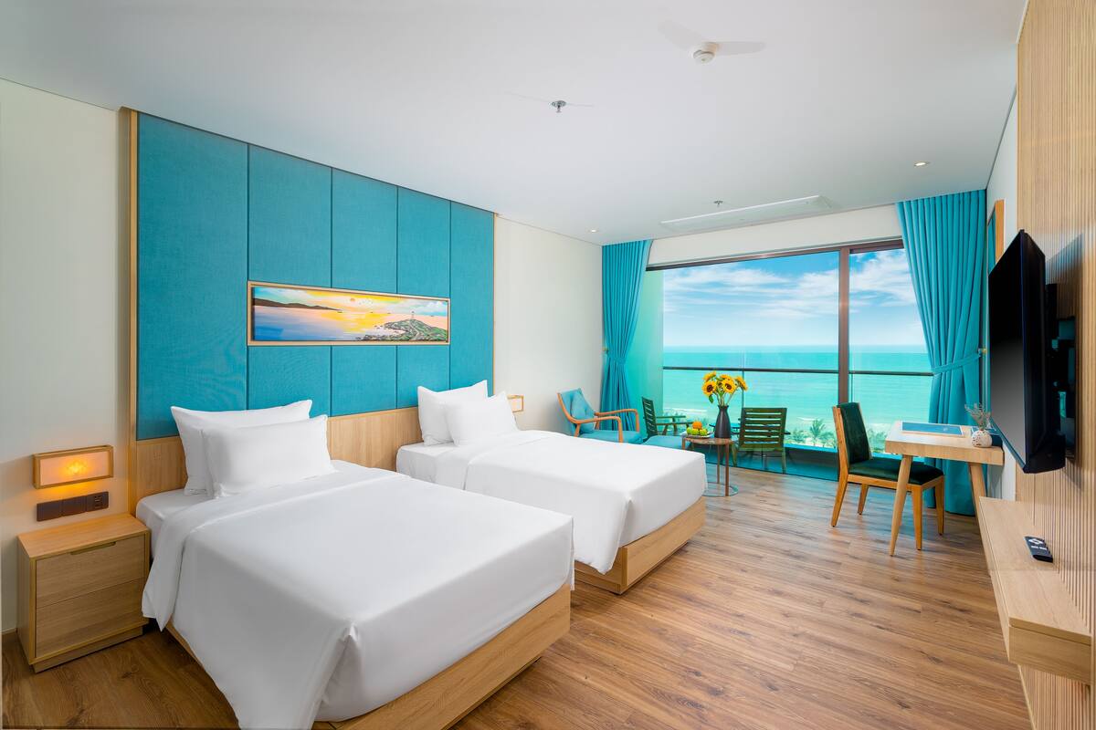 Ein Hotelzimmer mit zwei Betten, einem Fernseher, einem Schreibtisch und Meerblick.