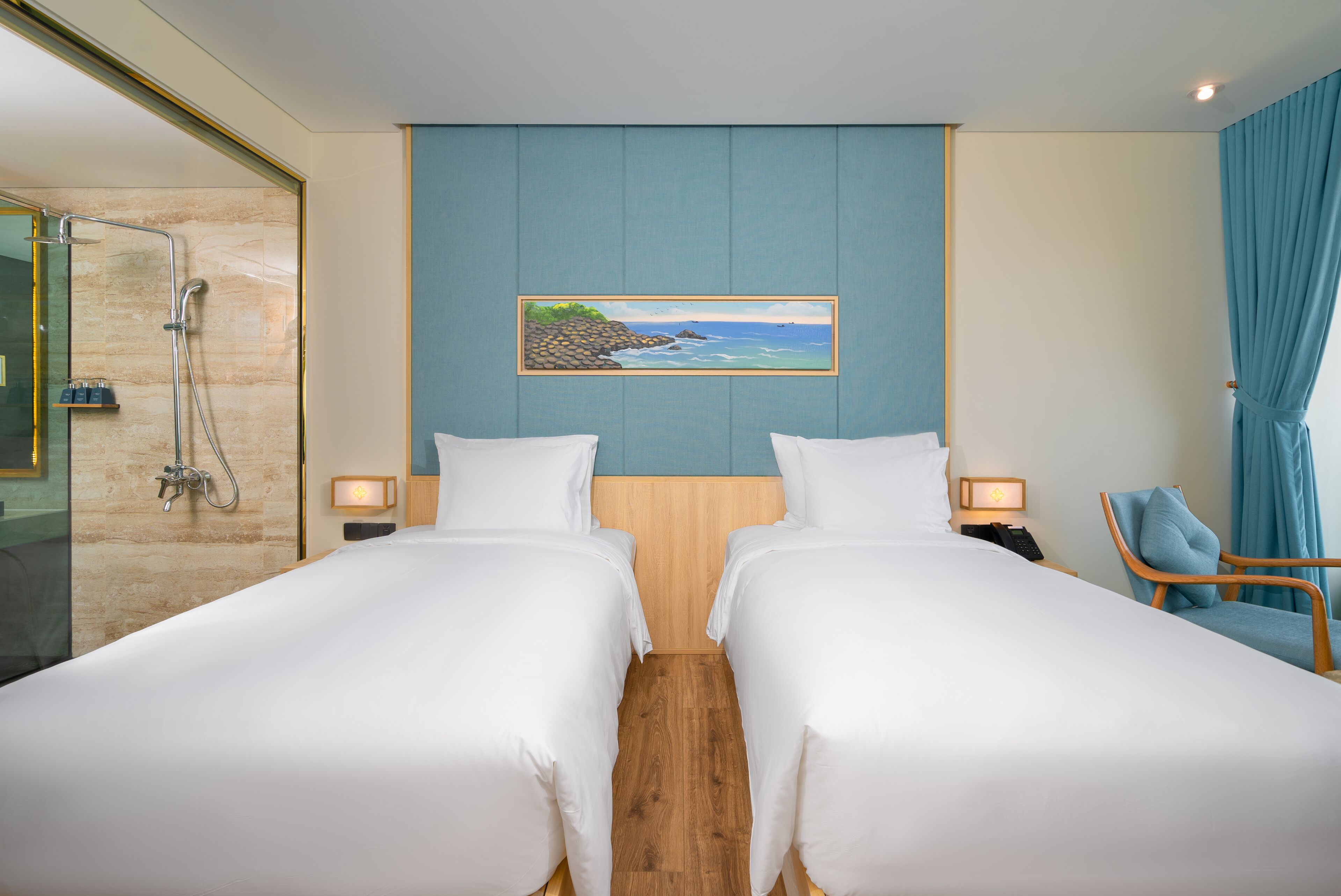 A hotel room with two beds, a shower, a painting of a beach, and a view of the sea.