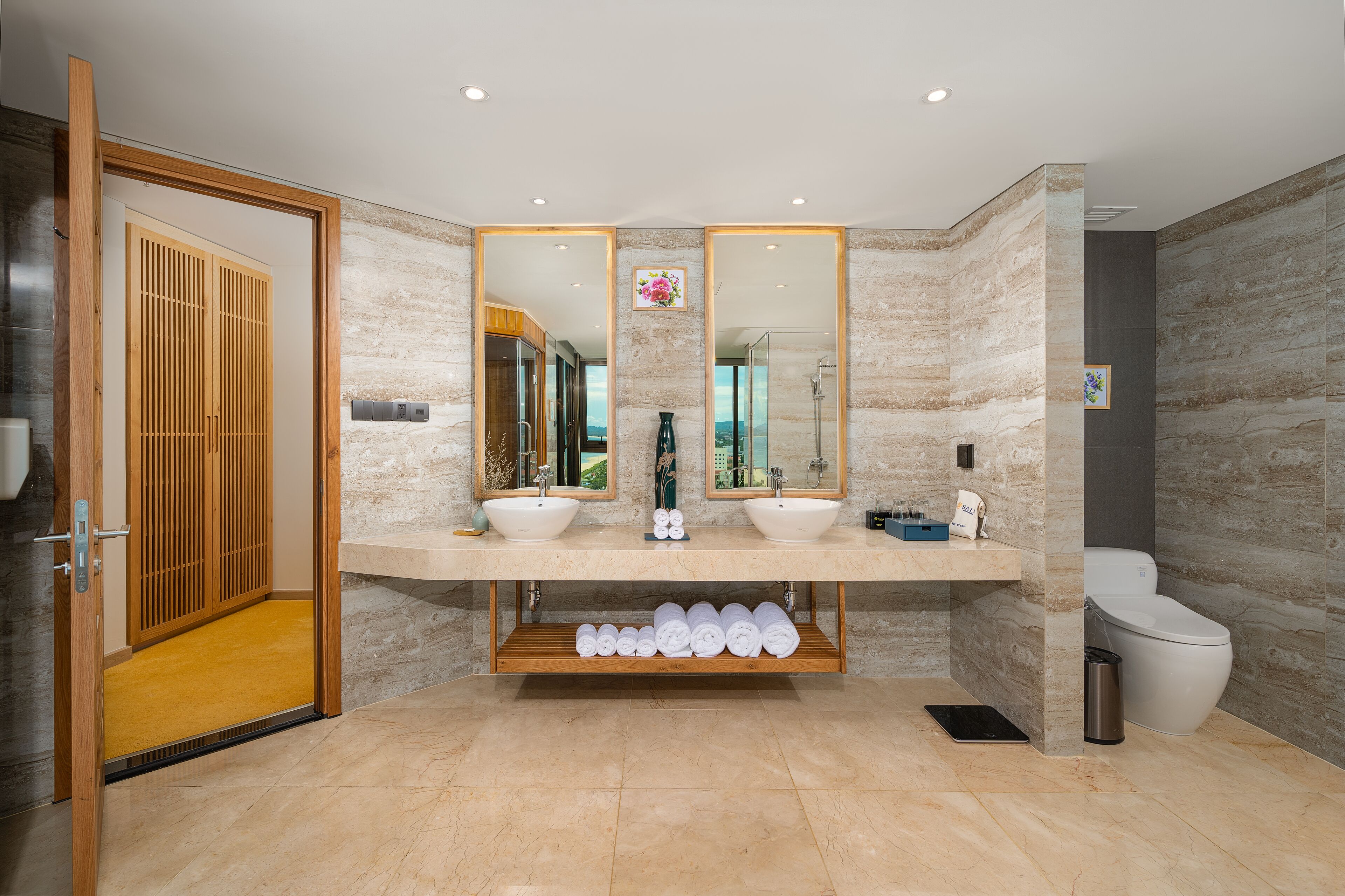 A modern bathroom with two sinks, a toilet, and a shower. There are wooden shelves under the sinks and a mirror above them.