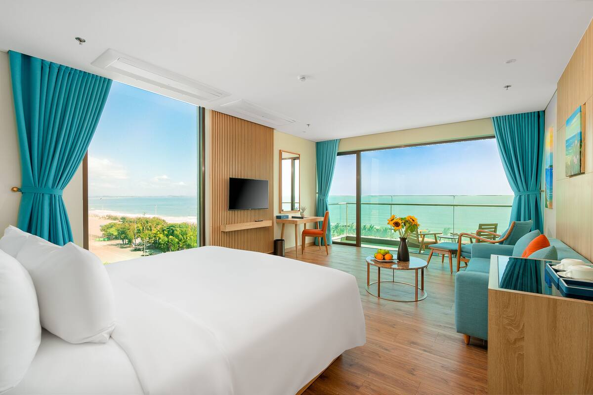 Ein Hotelzimmer mit einem großen Bett, einem Schreibtisch, einem Fernseher und einem Balkon mit Meerblick.