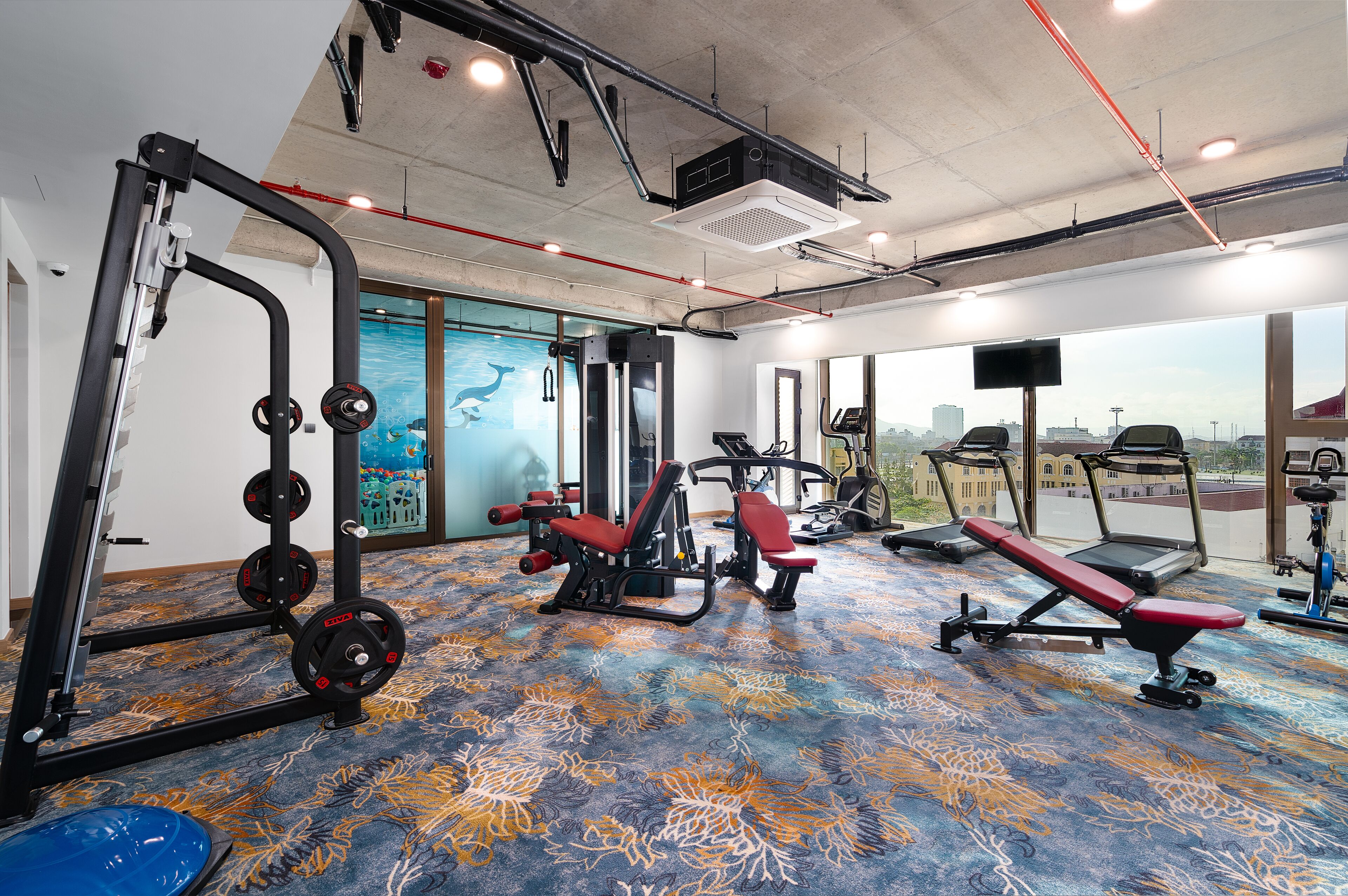 Een sportschool met diverse fitnessapparaten, een kamerbreed tapijt met dessin en grote ramen met uitzicht op de stad.