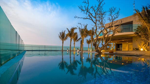 Un hôtel moderne doté d’une piscine à débordement dont l’eau reflète le ciel, de palmiers et offrant une vue sur un ciel dégagé.