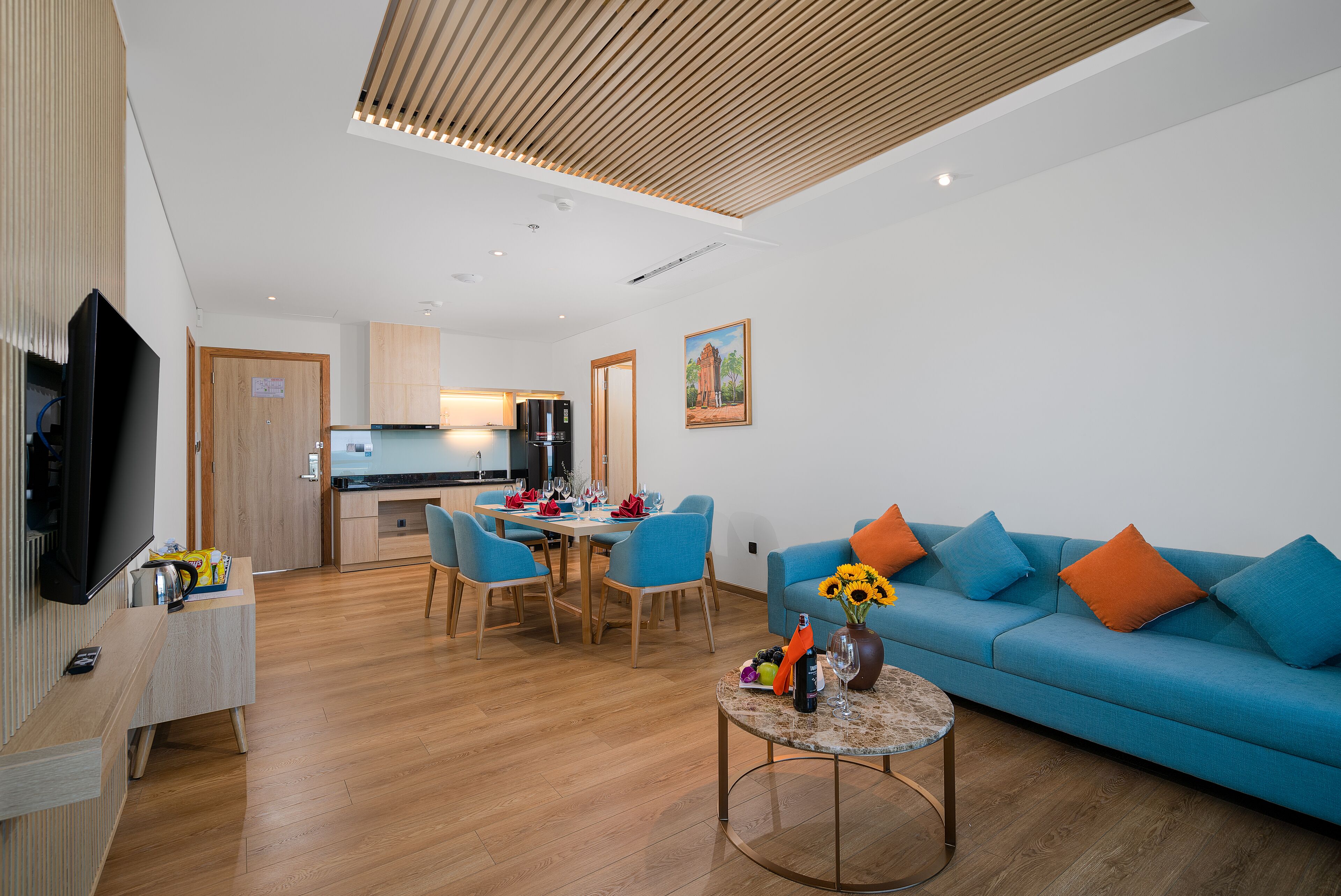 A modern living room with a blue sofa, wooden flooring, a dining area with blue chairs, a kitchen in the background, and a flat-screen TV on a stand.