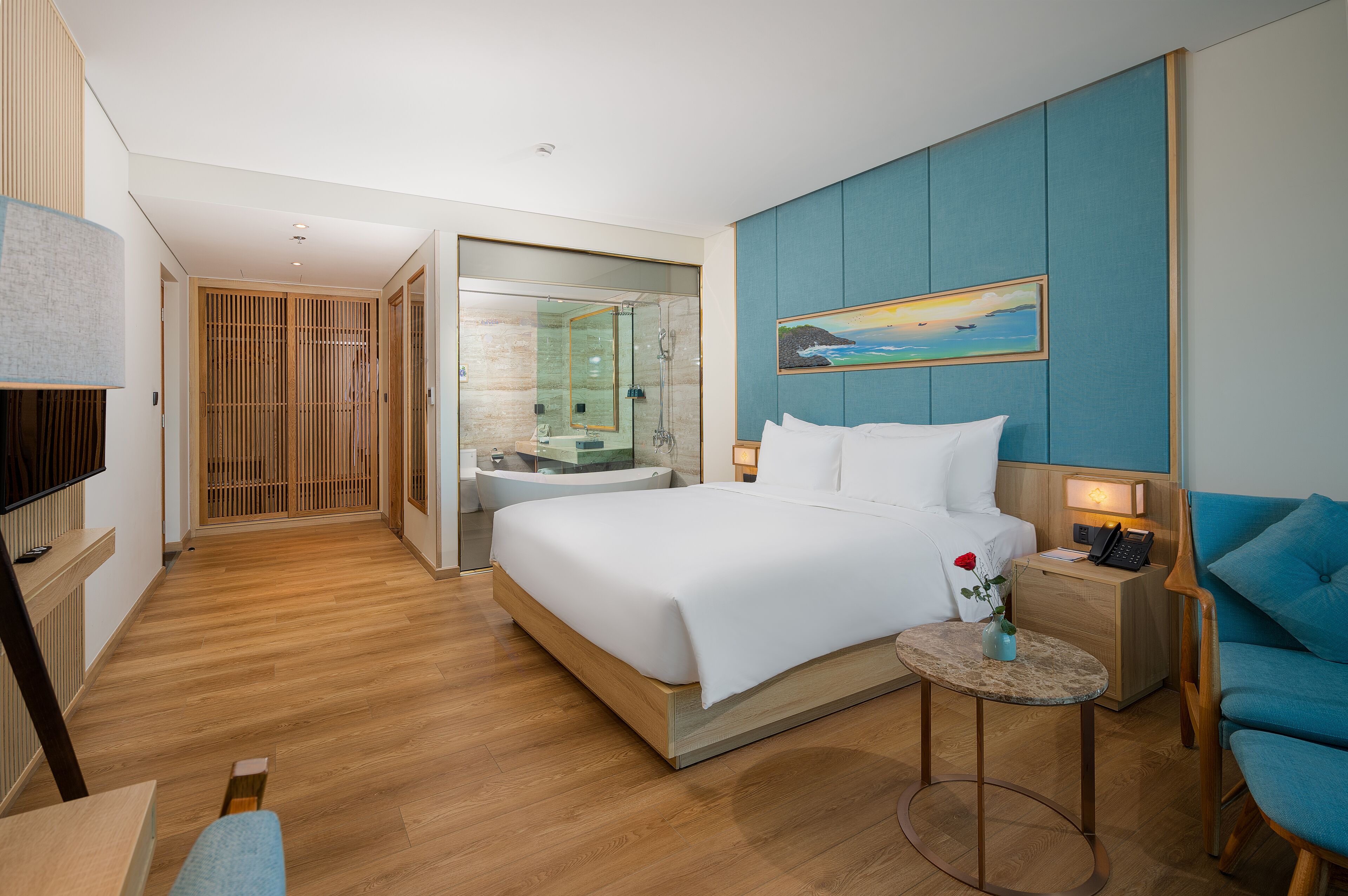 A modern hotel room with a large bed, a small bedside table with a lamp, a blue armchair, and a bathroom with a glass shower enclosure.