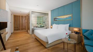 A modern hotel room with a large bed, a small bedside table with a lamp, a blue armchair, and a bathroom with a glass shower enclosure.