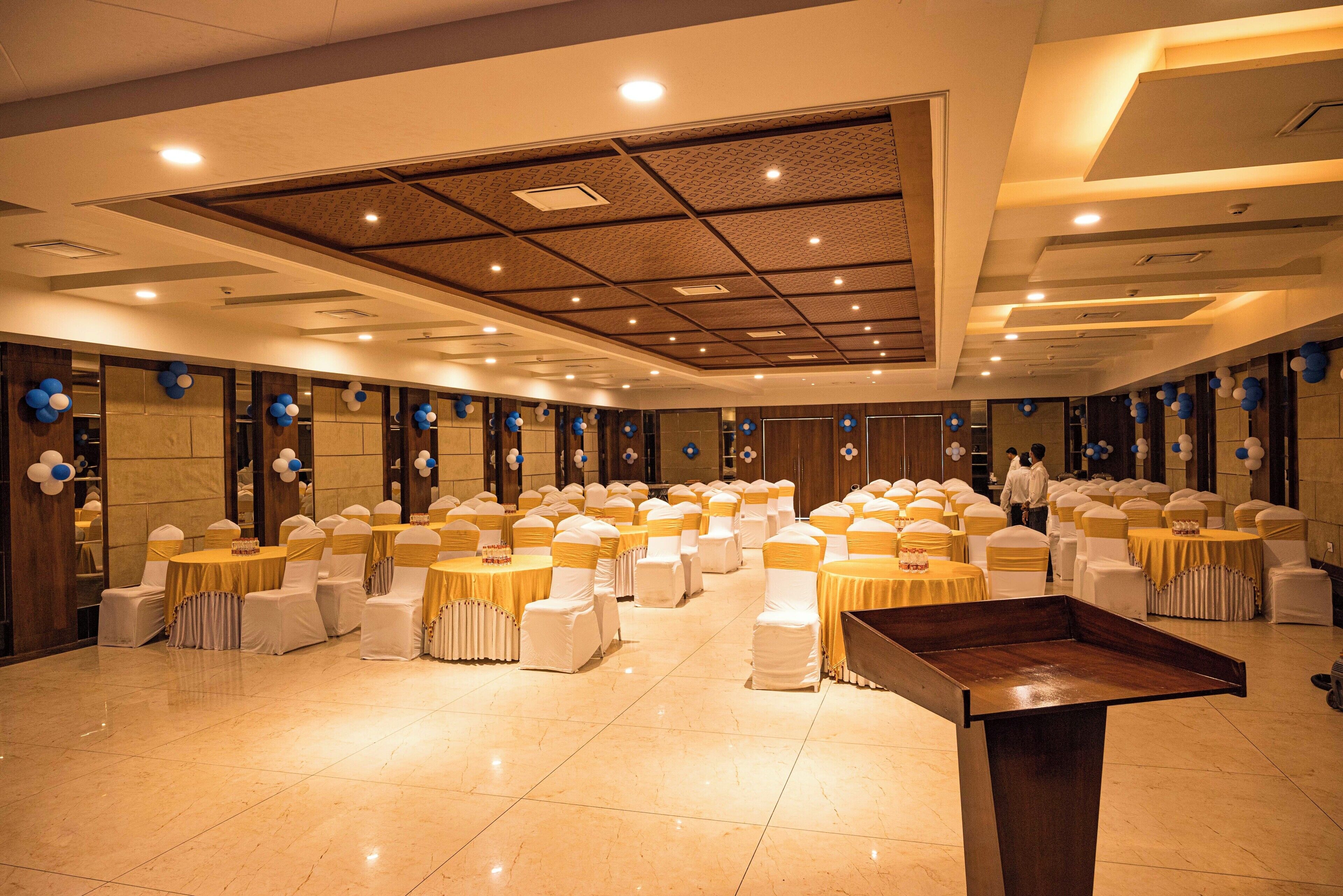 A spacious banquet hall with multiple round tables and chairs, a stage, and a high ceiling with recessed lighting.
