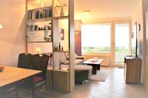 Una sala de estar con sofá, mesa de centro, mesa de comedor con sillas, estantería y televisor.