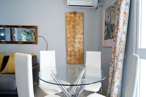 Une pièce moderne avec une table en verre, des chaises blanches, un canapé gris agrémenté de coussins jaunes, une grande œuvre d’art verticale et un tableau encadré accroché au mur.