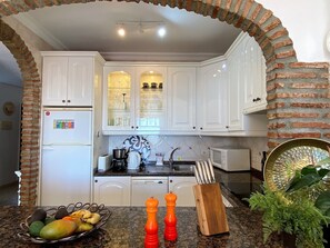 A kitchen with white cabinets, a brick archway, a refrigerator, a microwave, a sink, and a countertop with a fruit bowl and pepper shakers.