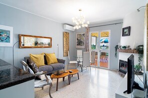 A modern living room with a grey sofa, a coffee table, and a television. There is a fireplace, a dining area with chairs, and a large window with curtains.
