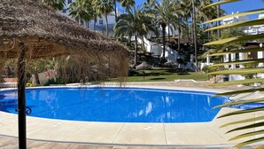 A clear blue swimming pool surrounded by palm trees and a thatched structure. - Oasis de Capistrano Rabat 27 by Casasol (Nerja)