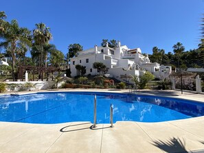 A swimming pool with a clear blue water, surrounded by a white building with balconies, palm trees, and a pergola.