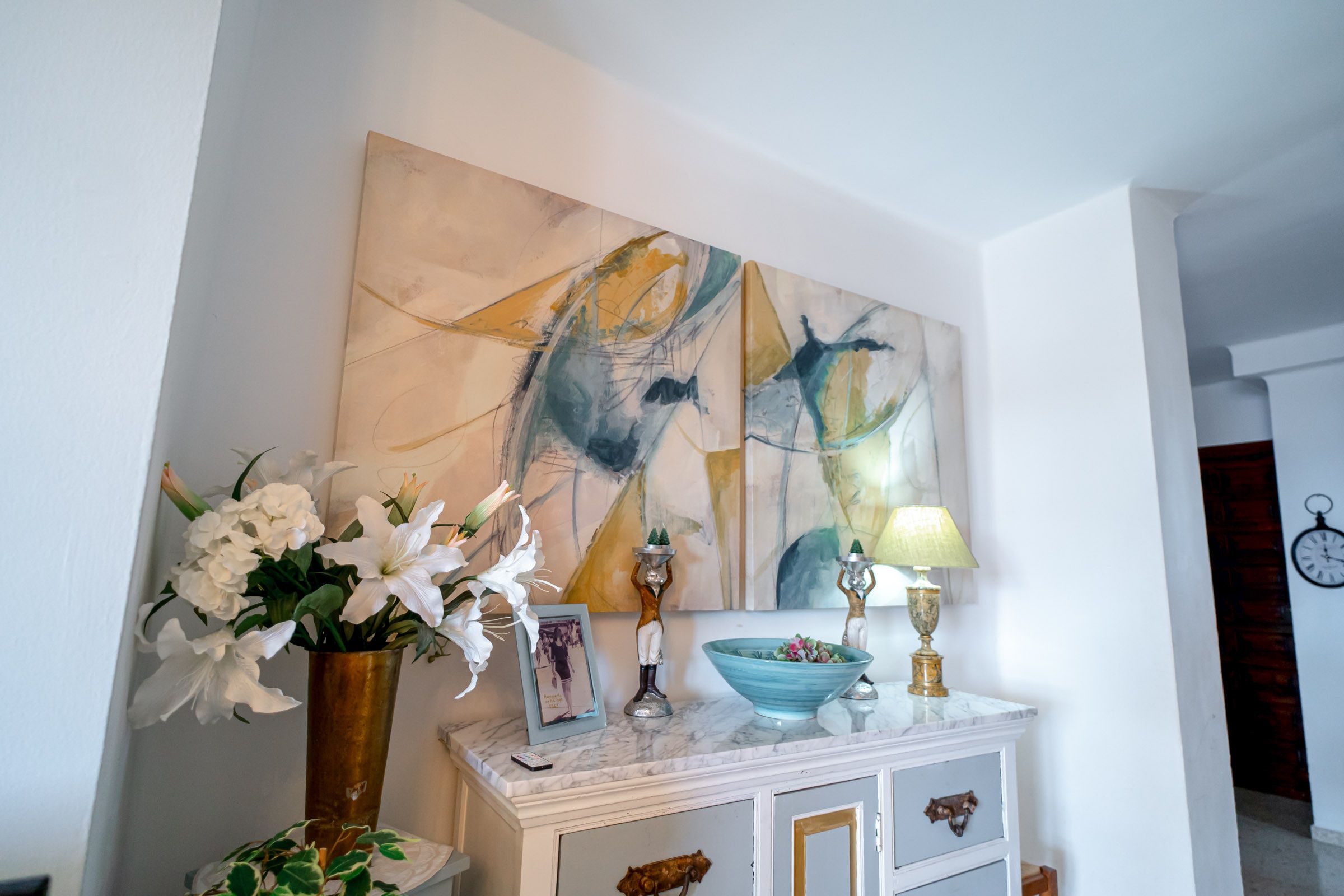 A room with a white console table, a vase with white flowers, a framed photograph, a teal bowl, and a wall-mounted abstract painting.