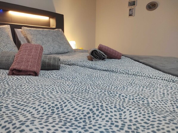 Una cama bien hecha con ropa de cama estampada, un cabecero con iluminación integrada y una lámpara de pared.