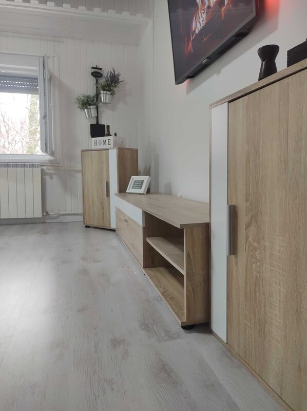 A modern living room with a wooden cabinet, a TV mounted on the wall, and a window with a view of trees. - Apartment Iskra (Plitvicka Jezera)