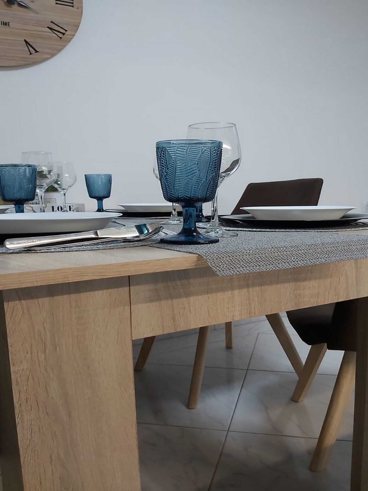 A dining table with a laptop, blue glassware, and a wooden wall clock.