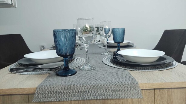 A dining table set with blue glassware and clear glasses, surrounded by plates and cutlery, with a wall clock above. - Apartment Iskra (Plitvicka Jezera)