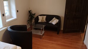 Una sala de estar con un sofá negro, una mesa de centro de vidrio, una planta en maceta y suelo de madera.