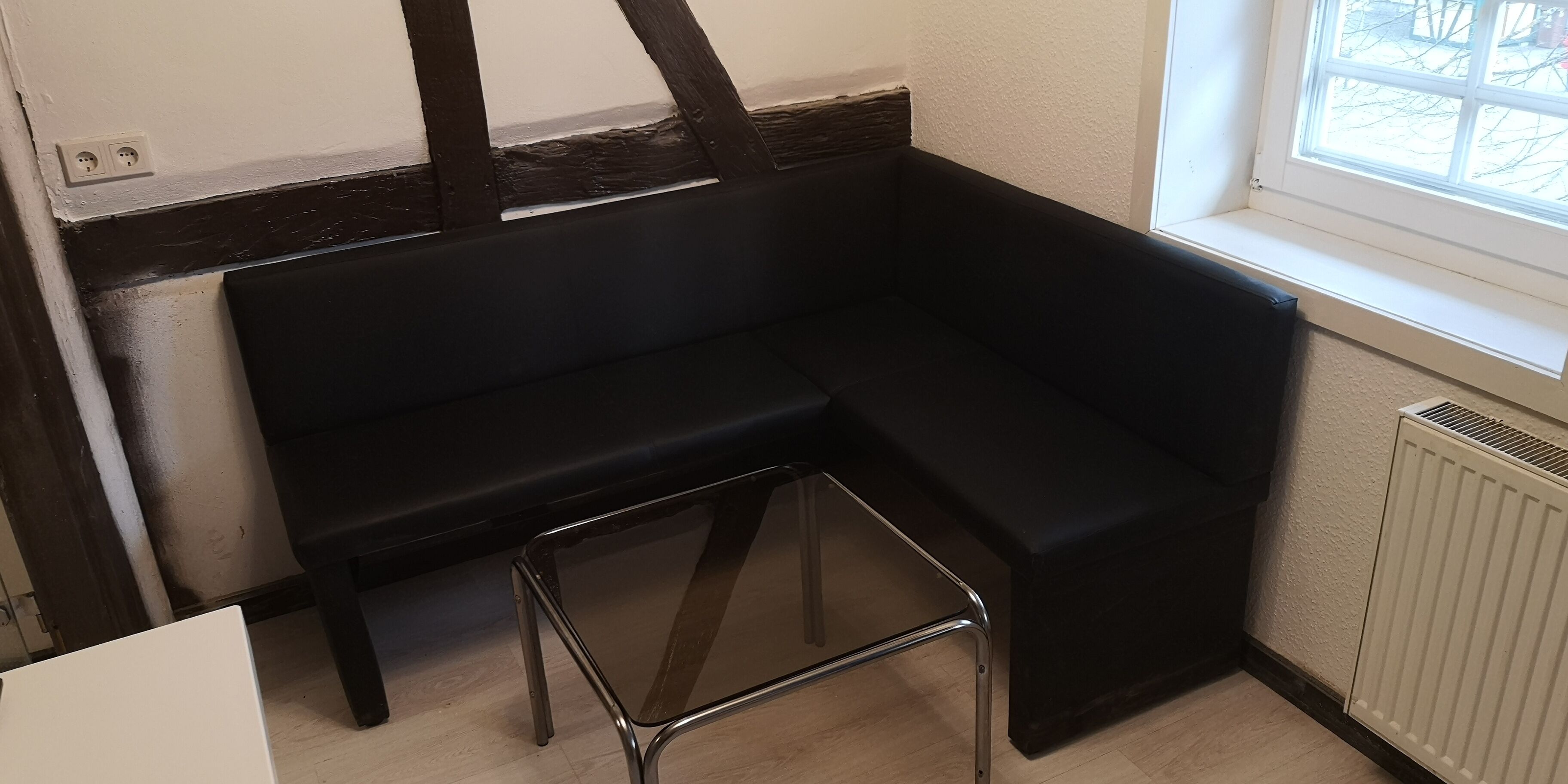 Une pièce d’angle avec un banc noir recouvert de tissu, un petit tabouret, un radiateur blanc et une fenêtre donnant sur l’extérieur.