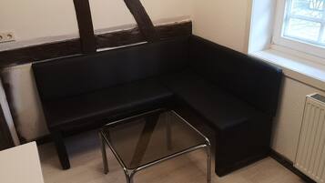 Une pièce d’angle avec un banc noir recouvert de tissu, un petit tabouret, un radiateur blanc et une fenêtre donnant sur l’extérieur.