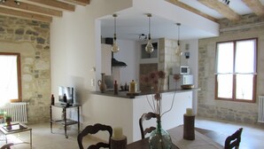 A dining area with a wooden table, chairs, a vase with flowers, and a kitchen in the background. - Le clos des oiseaux is a winery located in Aimargues. (Aimargues)